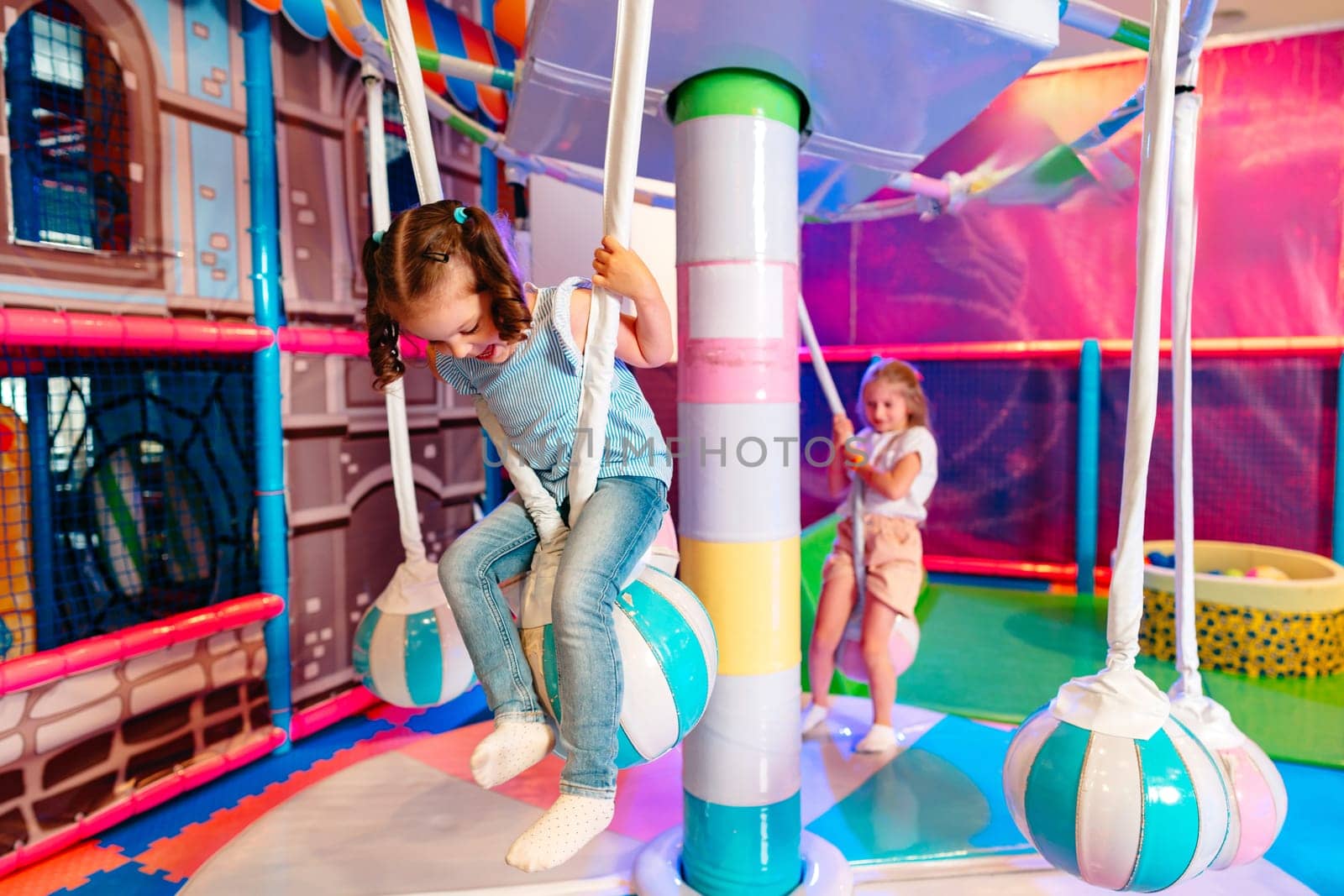 A cheerful young girl with a ponytail is having fun climbing on colorful play equipment at an indoor playground. She is wearing a sleeveless top and pants, while another child is visible in the background, adding to the lively atmosphere.