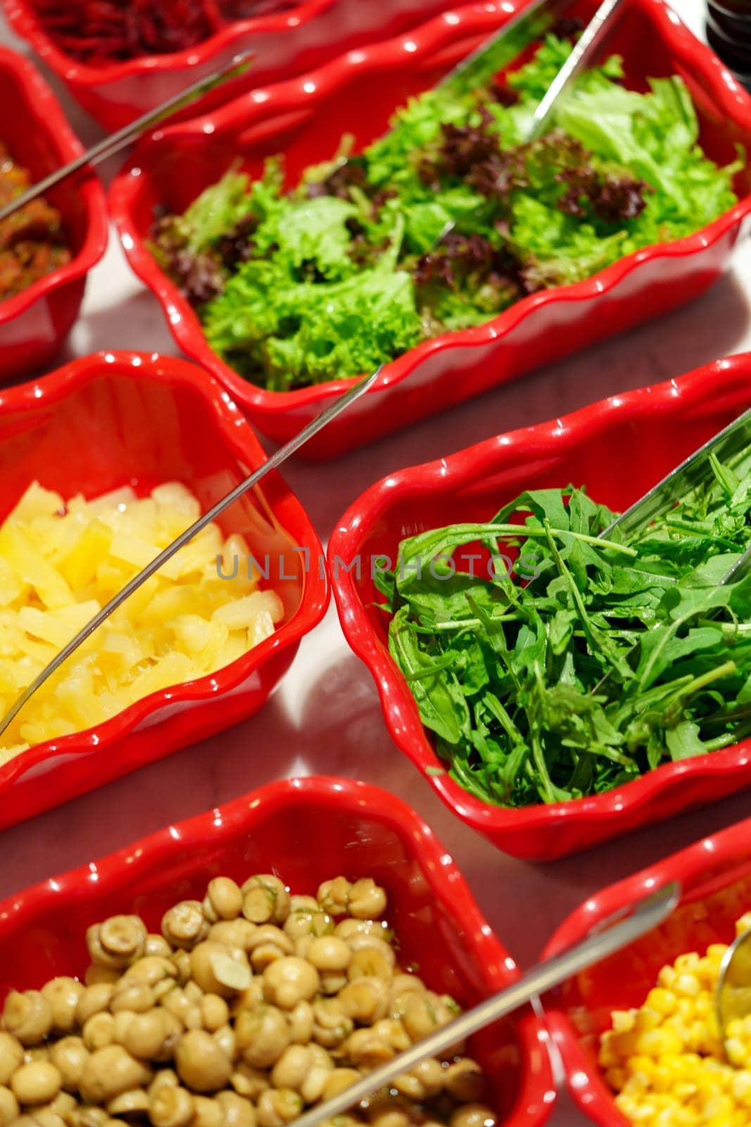 A variety of fresh salad ingredients are neatly presented in red dishes forming a vibrant buffet line. The bowls are filled with crisp greens, sweetcorn, and other tasty vegetables, inviting diners to create their own salad masterpiece. Each container is equipped with serving tongs or spoons for convenience, and condiments are provided along the table to enhance the flavors.