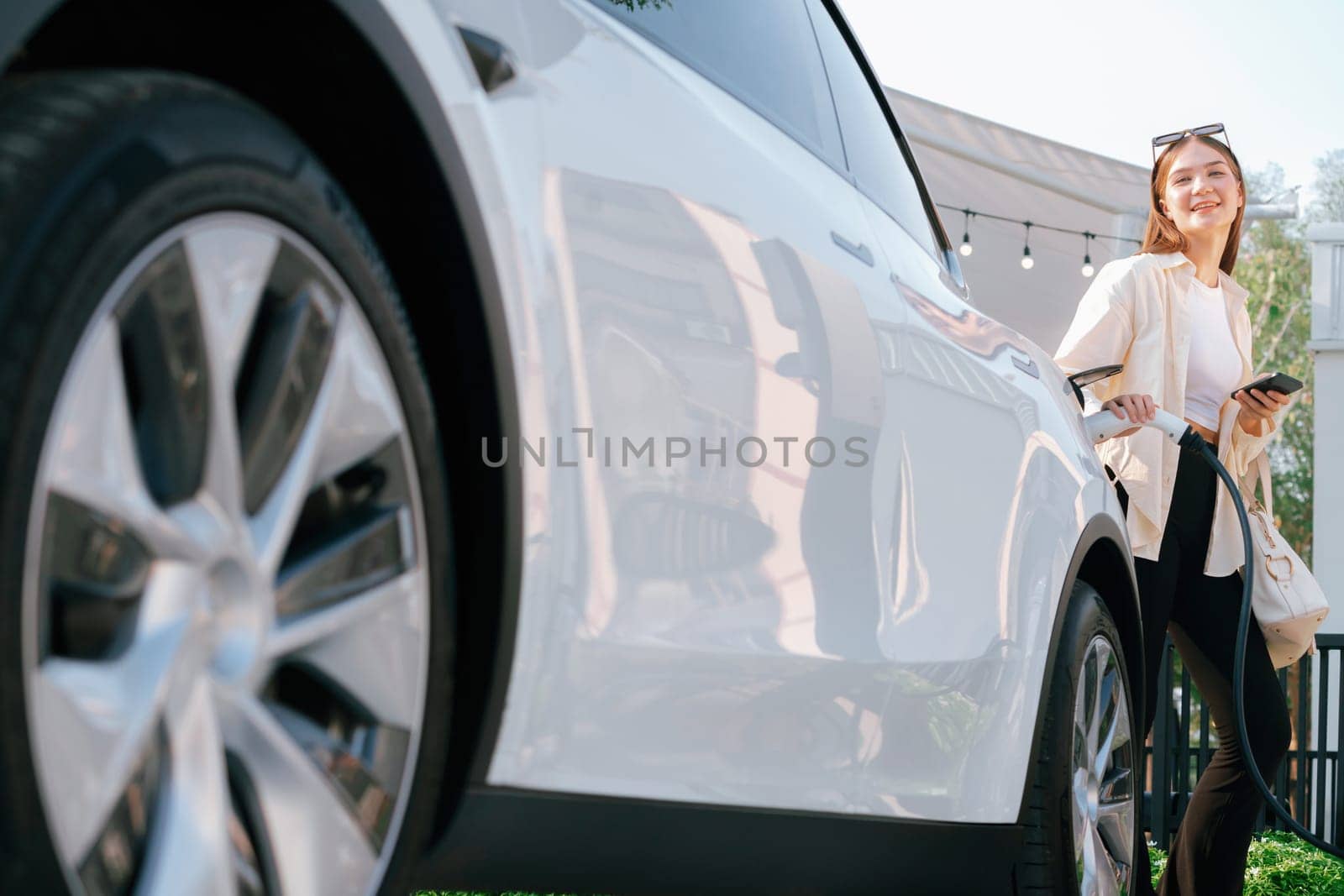 Sustainable urban commute with young woman pay electricity for EV electric car recharging at outdoor cafe in springtime garden, green city sustainability and environmental friendly EV car. Expedient