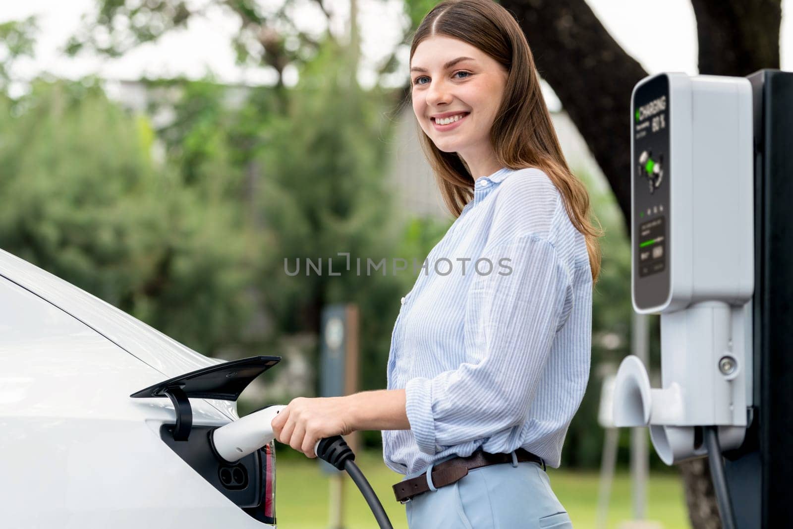 Young woman recharge EV electric vehicle's battery from EV charging station in outdoor green city park scenic. Eco friendly urban transport and commute with eco friendly EV car travel. Exalt