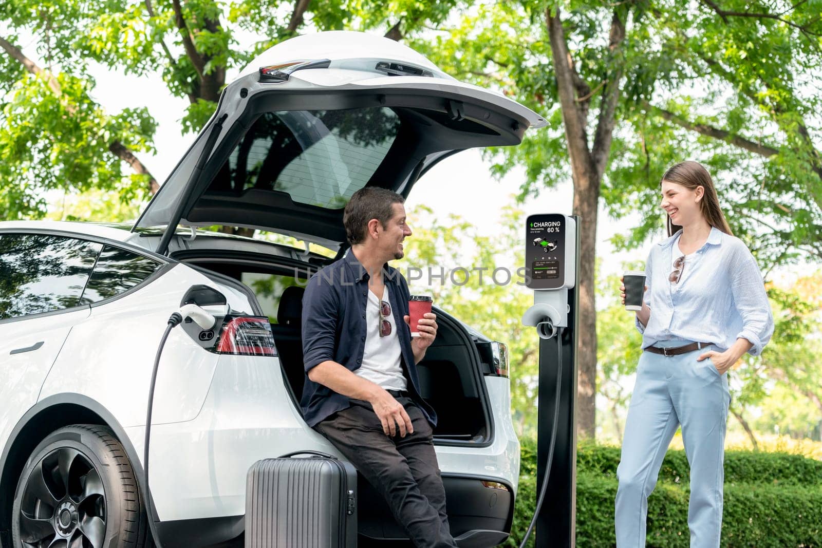 Lovely young couple drinking coffee while recharging battery for electric car during road trip travel EV car in natural forest or national park. Eco friendly travel during vacation and holiday. Exalt