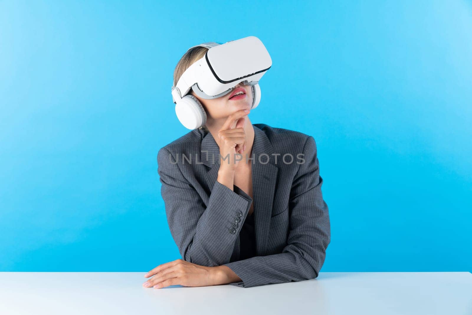 Smart project manager wearing VR glasses while sitting and thinking about marketing plan. Skilled business woman using visual reality goggles and sitting at blue background. Innovation. Contraption.