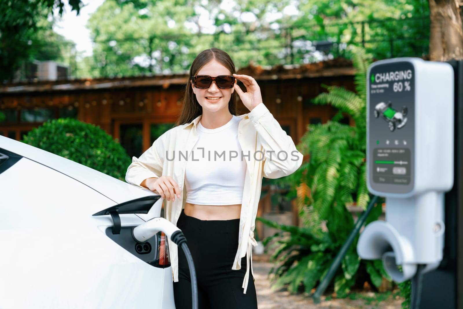 Young woman and sustainable urban commute with EV electric car recharging at outdoor cafe in springtime garden, green city sustainability and environmental friendly EV car. Expedient