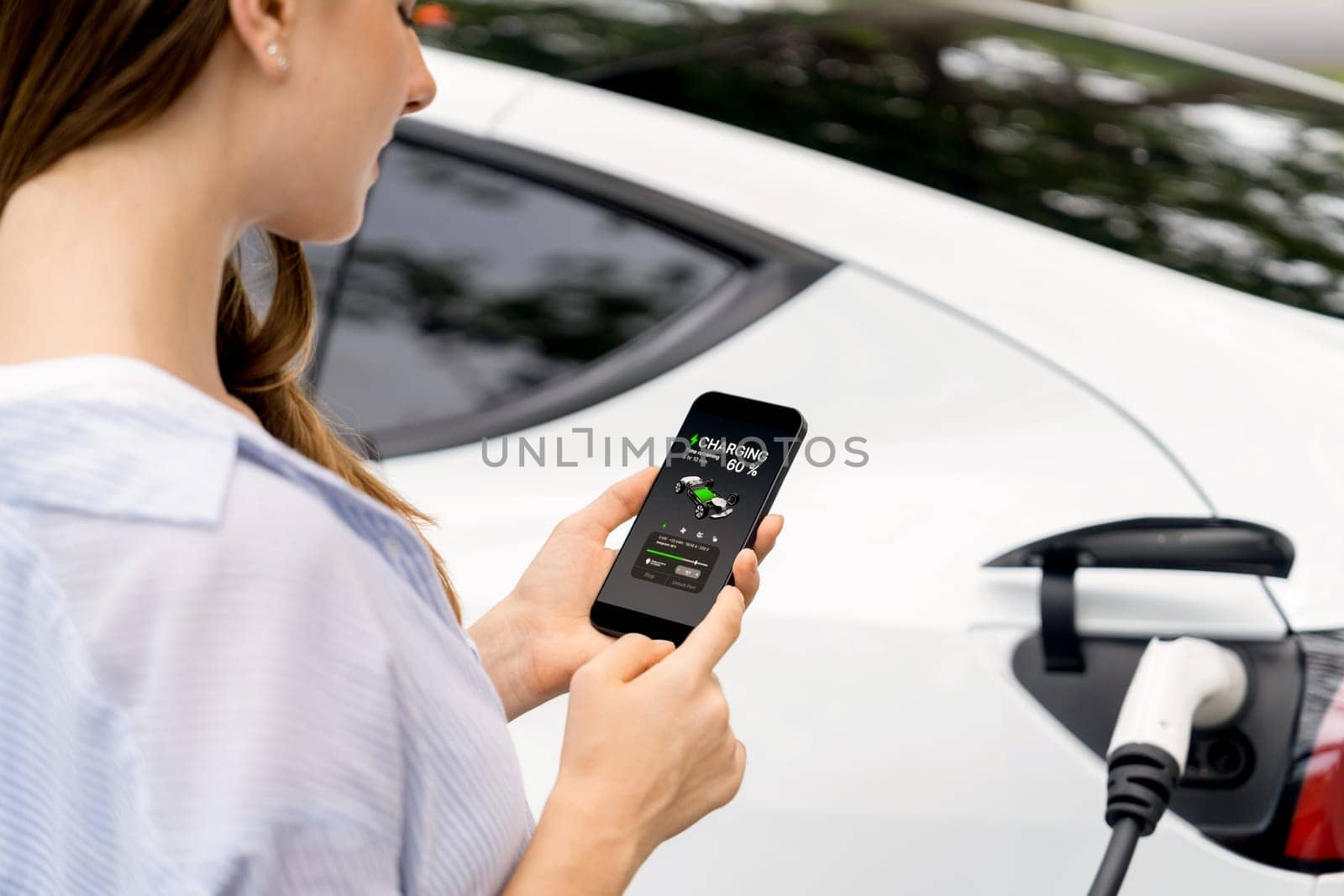 Young woman using smartphone online banking application to pay for electric car battery charging from EV charging station during vacation holiday road trip at national park or summer forest. Exalt