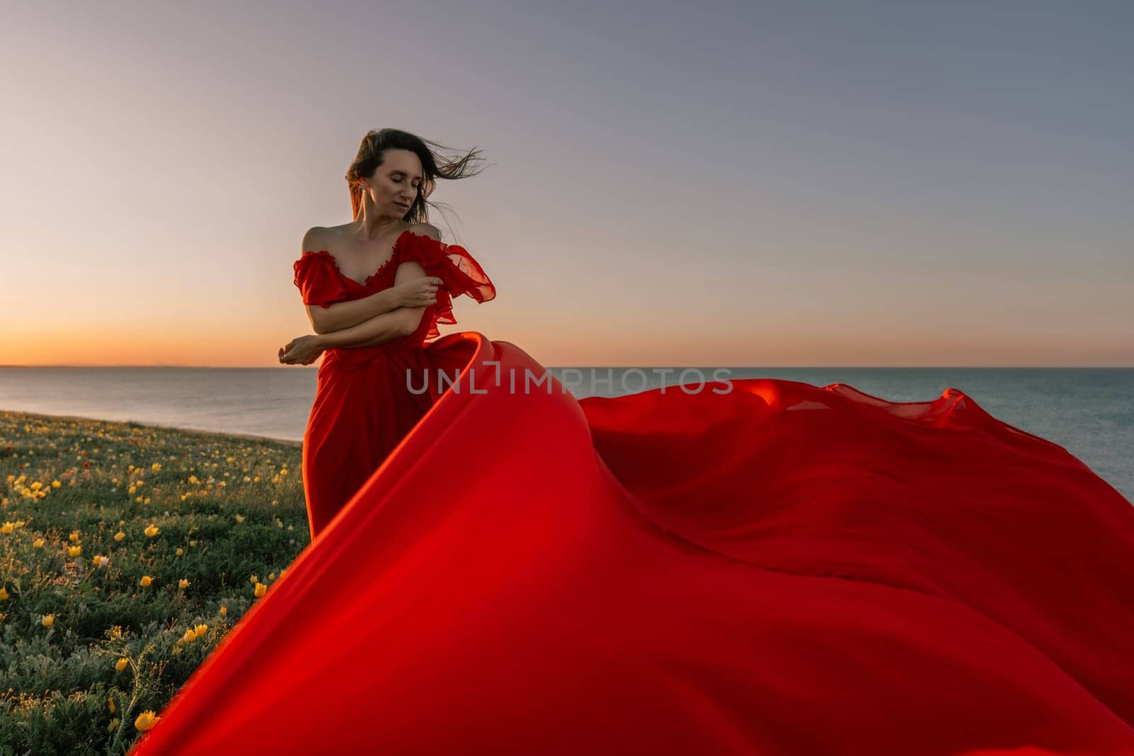 woman red dress standing grassy hillside. The sun is setting in the background, casting a warm glow over the scene. The woman is enjoying the beautiful view and the peaceful atmosphere