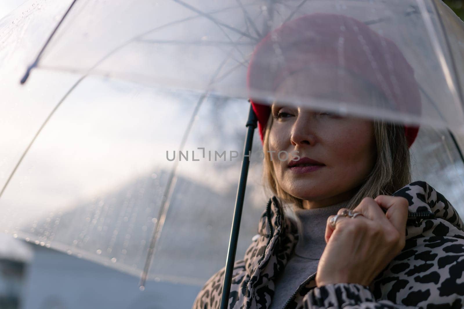 A woman wearing a red hat and a black and white jacket is holding a clear umbrella. The umbrella is open, and the woman is looking up at the sky. Concept of solitude and contemplation