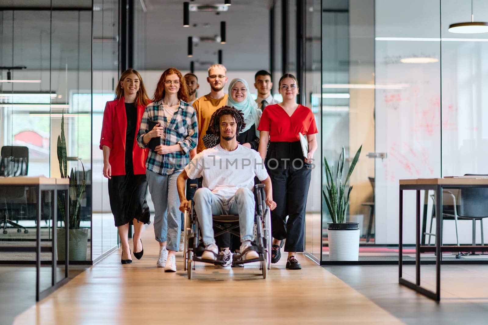 A diverse group of young business people walking a corridor in the glass-enclosed office of a modern startup, including a person in a wheelchair and a woman wearing a hijab by dotshock