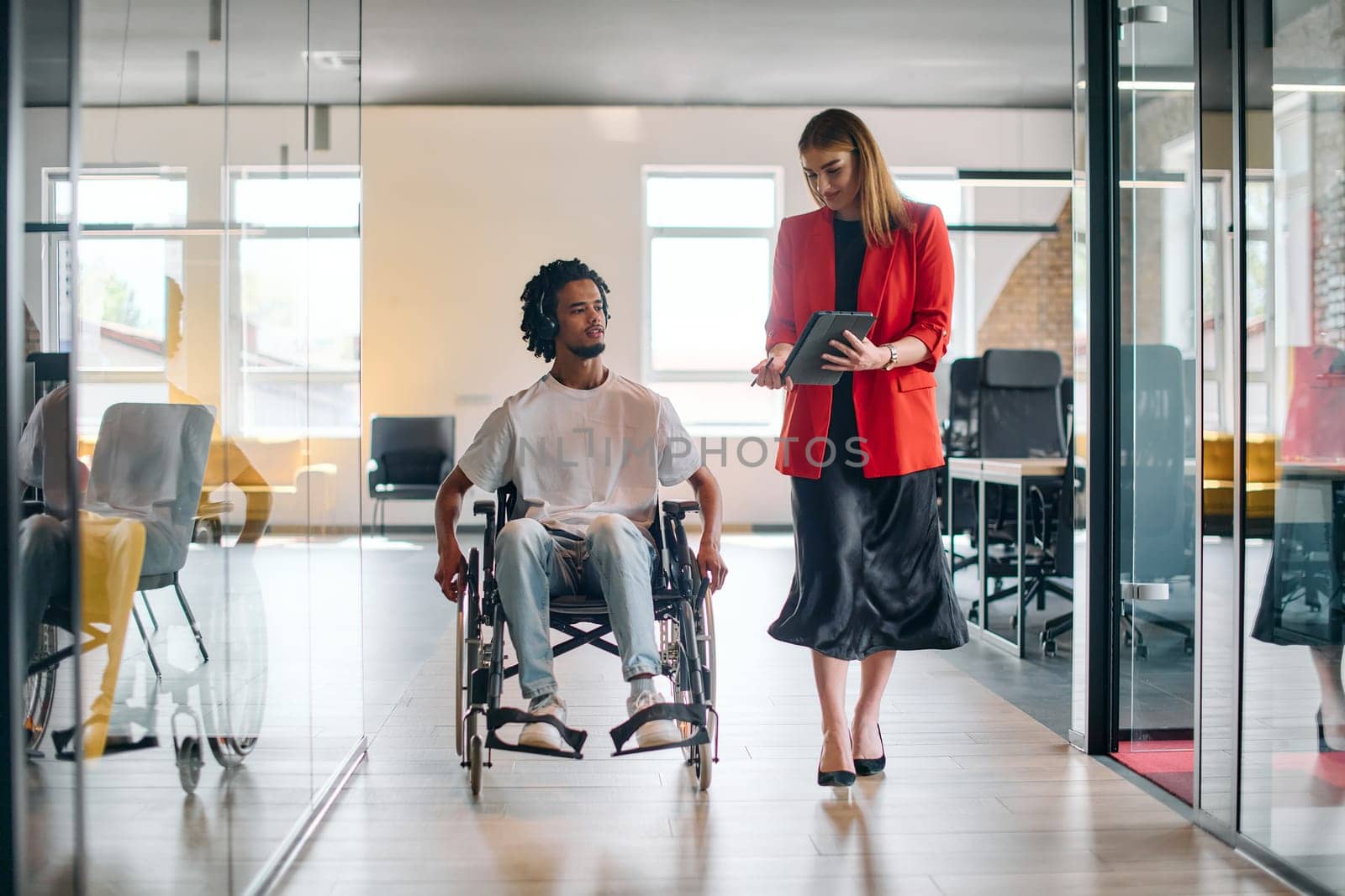 A business leader with her colleague, an African-American businessman who is a disabled person, pass by their colleagues who work in modern offices.