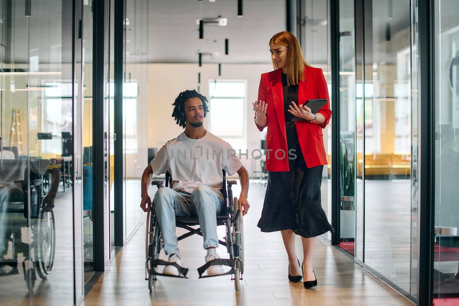 A business leader with her colleague, an African-American businessman who is a disabled person, pass by their colleagues who work in modern offices.