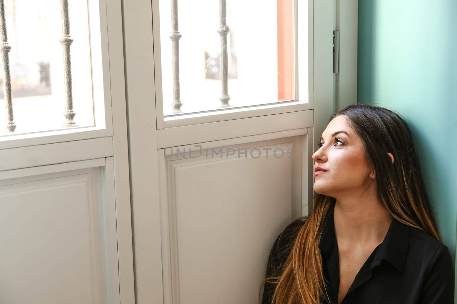 Young model, dressed in a black shirt, looking out by the window, illuminated only by natural light.