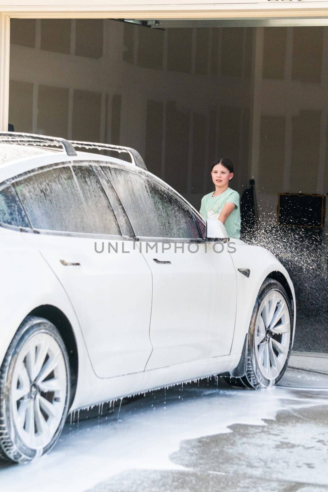 Denver, Colorado, USA-September 1, 2023-A young girl enthusiastically assists in washing the family's electric car in their suburban driveway.