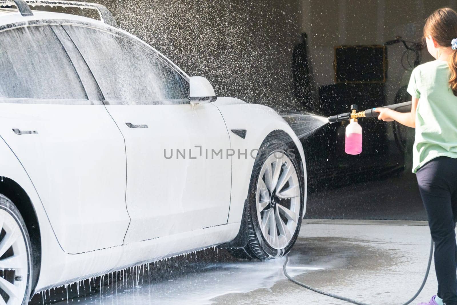 Denver, Colorado, USA-September 1, 2023-A young girl enthusiastically assists in washing the family's electric car in their suburban driveway.