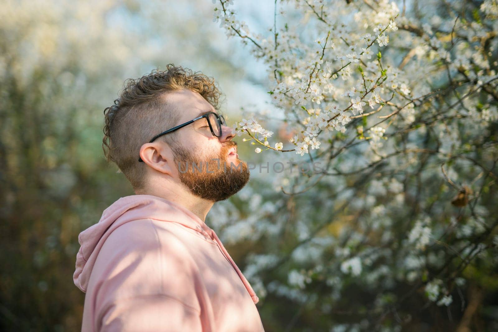 Portrait of curly millennial man inhales the fragrance of spring flowers of blooming jasmine or cherry tree. Spring time concept. Copy space by Satura86