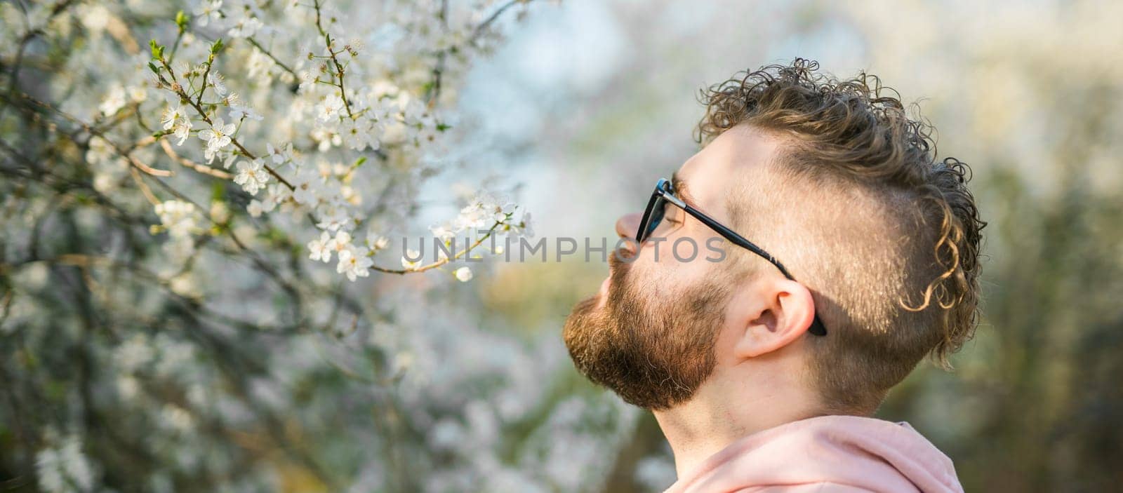 Banner portrait of curly millennial man inhales the fragrance of spring flowers of blooming jasmine or cherry tree. Spring time concept. Copy space by Satura86