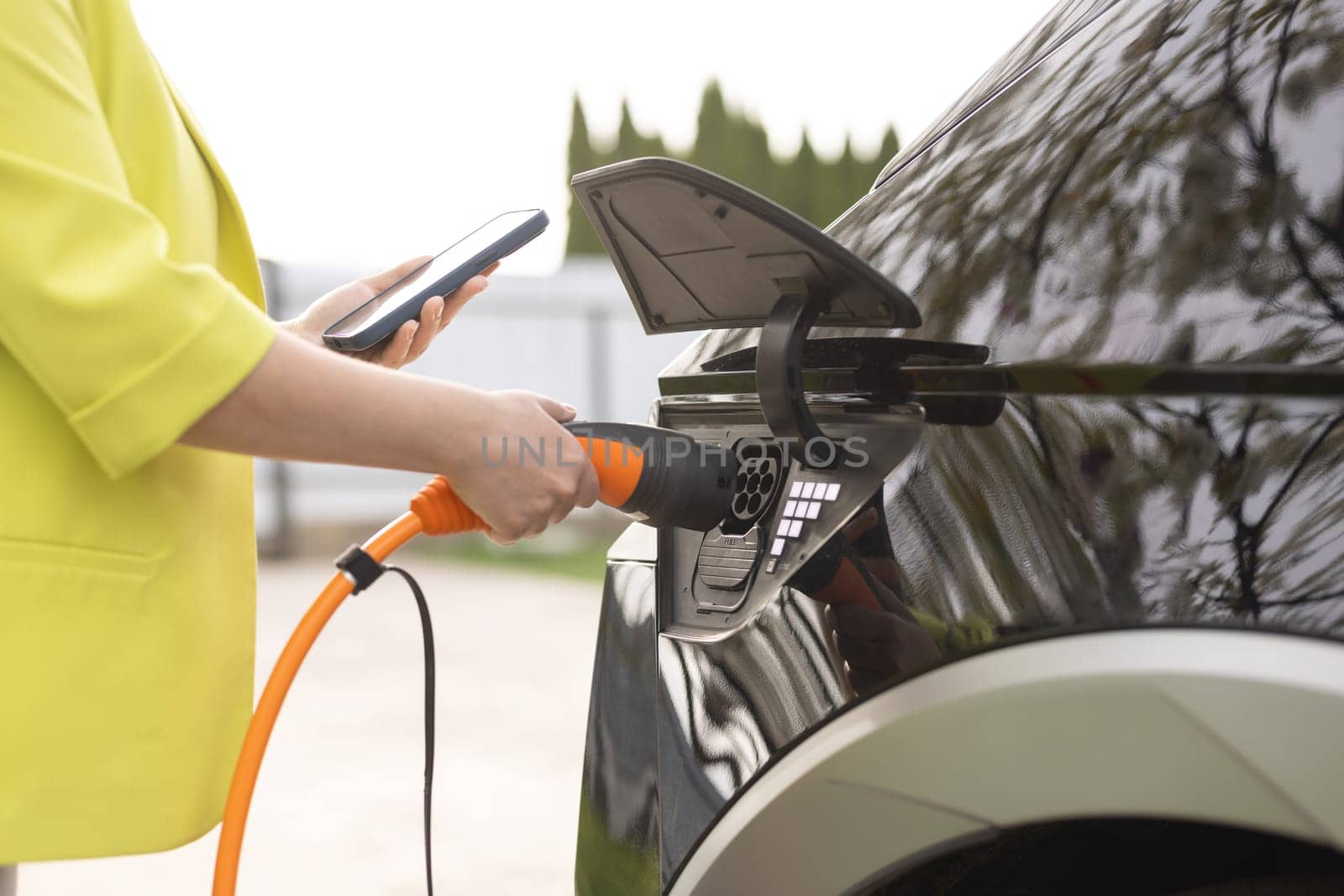 Woman connects an electric car to the charger and adjusts the process of charging the car battery using a cell phone smart phone. Girl plugs power cable to charge electric car in parking lot. by uflypro