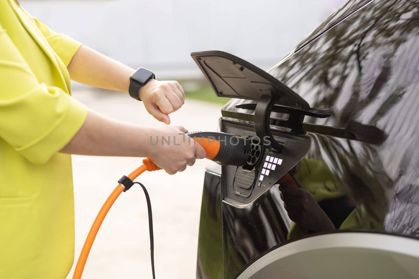 Female plugging an electric car or EV at electric charging station. Woman hands attaching power cable supply to charge electric or EV car using app on wearable smart watch.