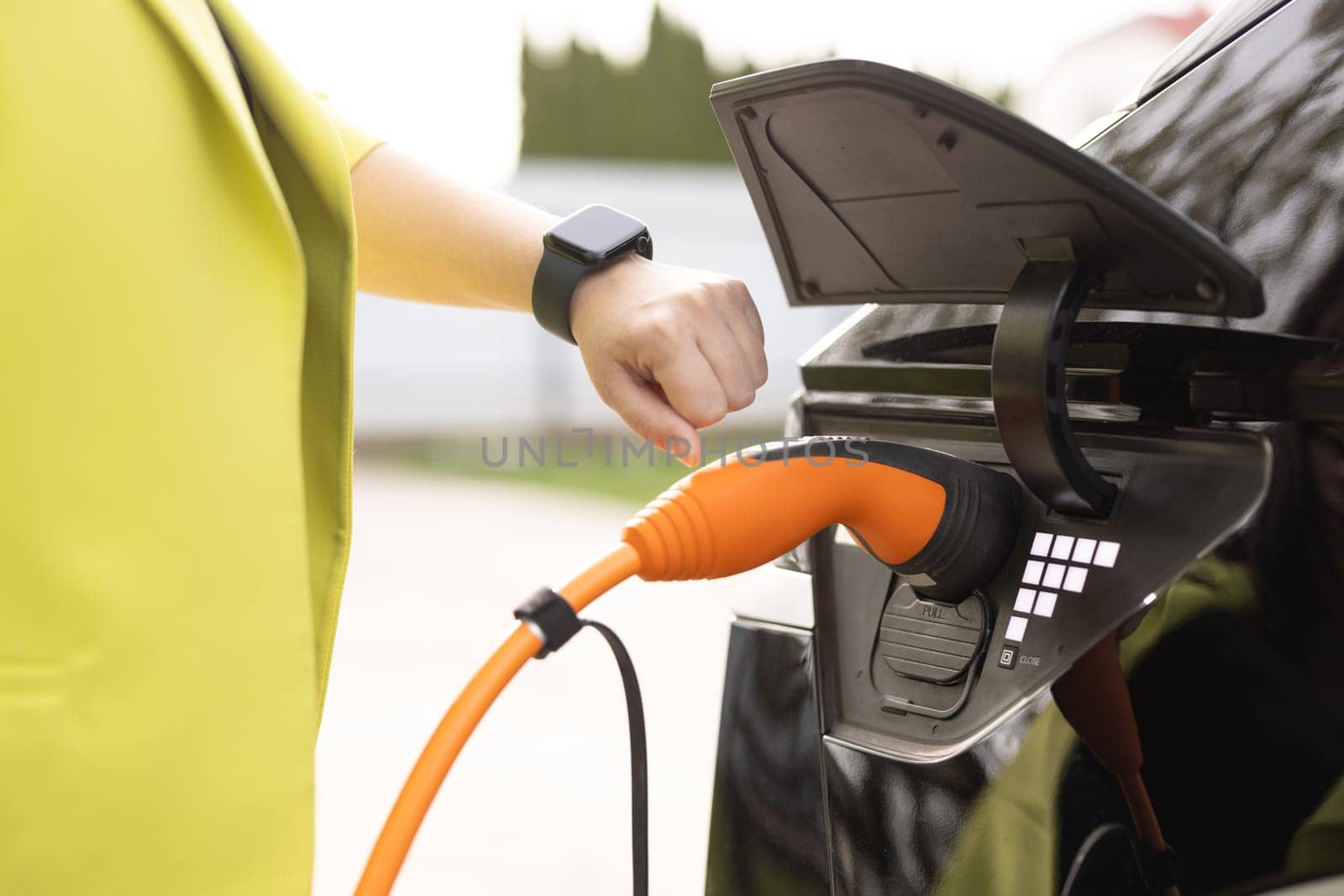 Woman hands attaching power cable supply to charge electric or EV car using app on wearable smart watch. Female plugging an electric car or EV at electric charging station. by uflypro