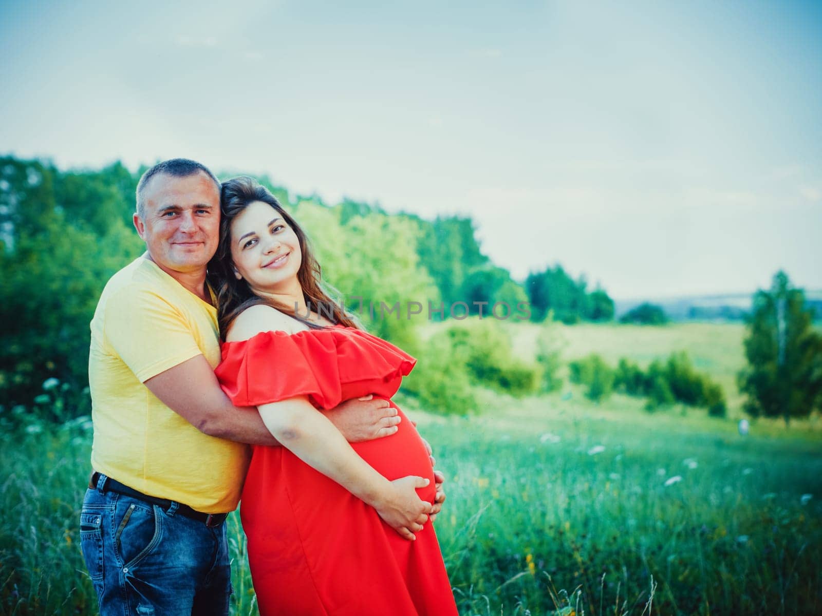 Smiling beautiful pregnant woman in red dress and her husband with hands on belly outdoors. Man embraces from behind belly pregnant wife and smiling