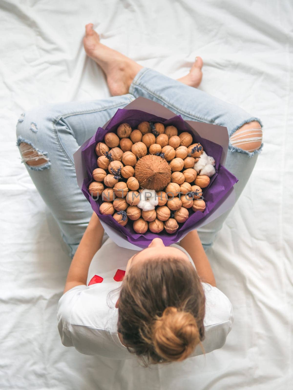 Young woman sitting on bed with coconut and walnuts eating bouquet in hands. View from above on young hipster woman in torn jeans at knee with unusual bouquet. Vertical. Copy space.