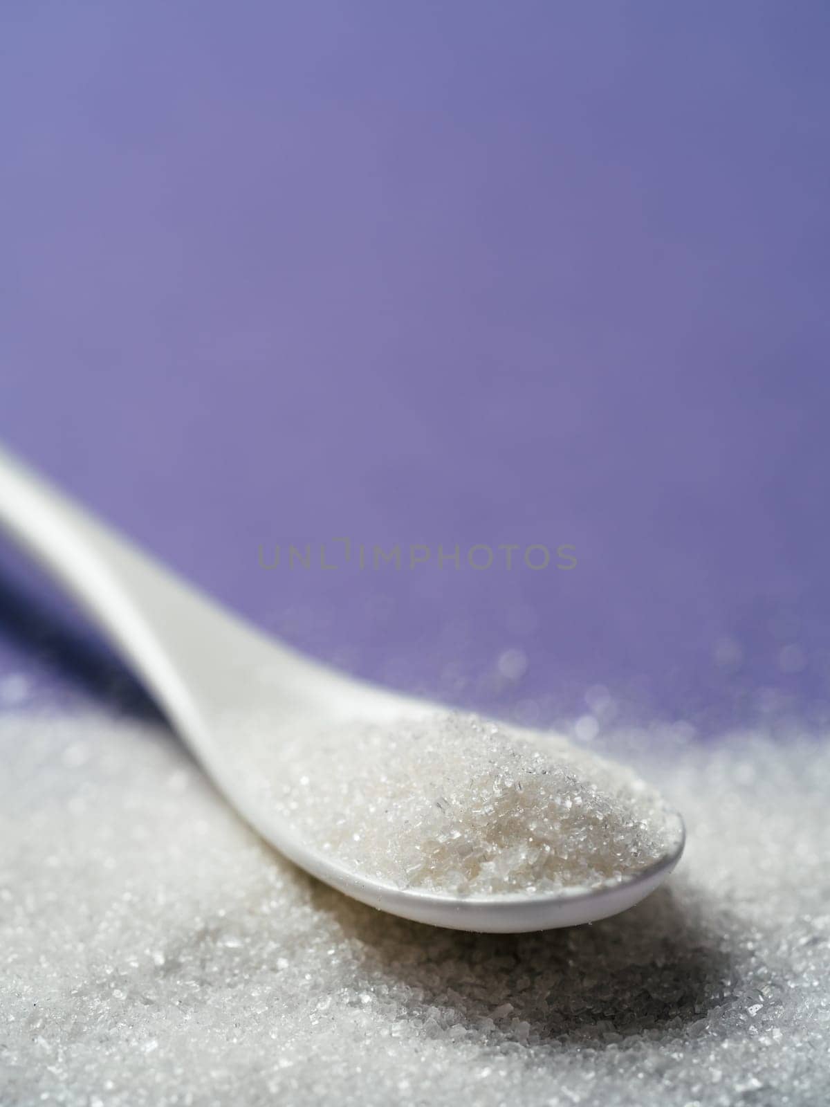 Sugar in white ceramic spoon on lilac background. Spoon with white sugar on violet background, closeup. Shallow DOF. Copy space for text. Vertical.