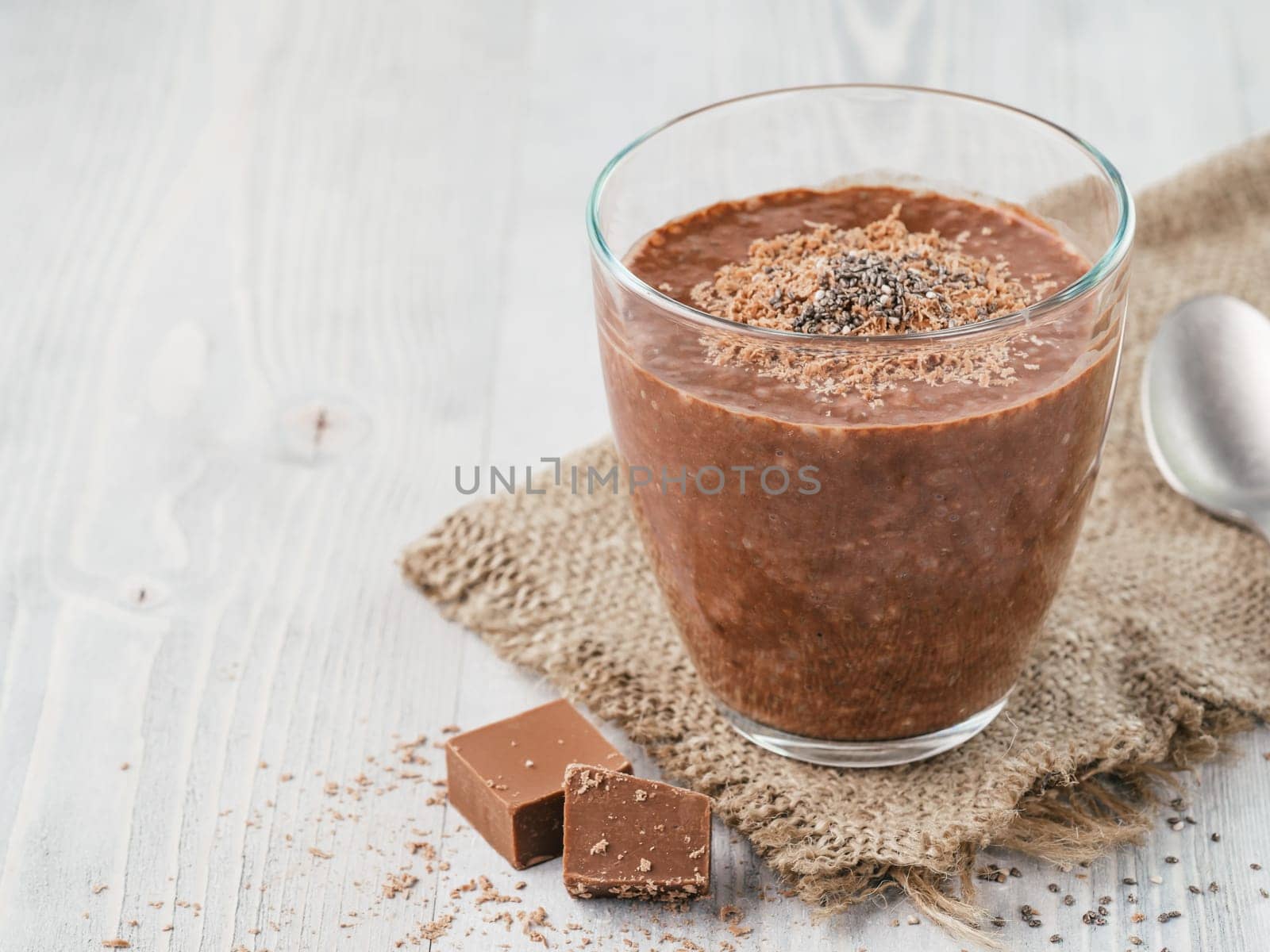 Chocolate chia pudding in glass and chocolate cubes slice with rustic napkin on gray wooden table. Healthy vegan breakfast with copy space.