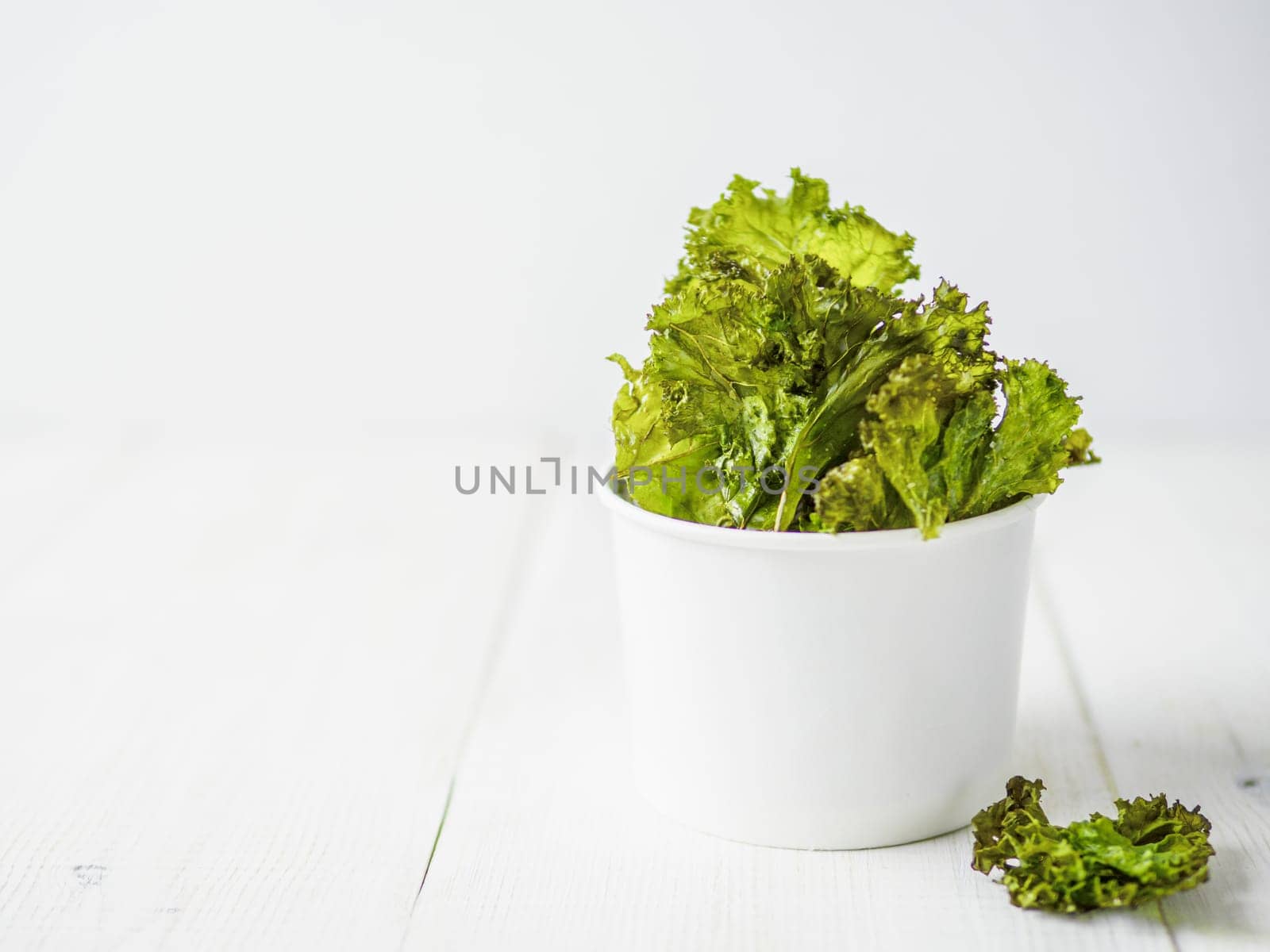 Kale Chips with salt in paper cup. Homemade healthy snack for low carb, keto, low calorie diet. White wooden background. Ready-to-eat kale chips, copy space for text. Banner