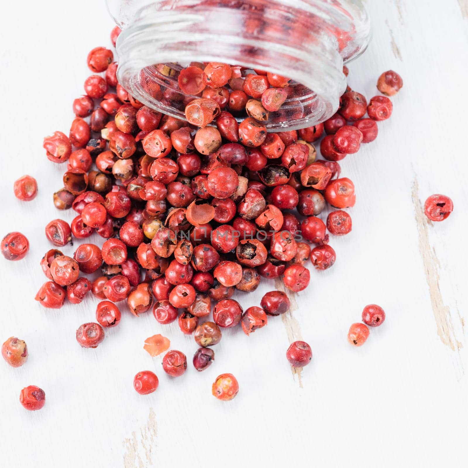 Dried pink pepper berries poured from glass jar on white wooden cutting board. Close up view of pink peppercorn poured on vintage wooden table. Copy space. Top view or flat-lay.