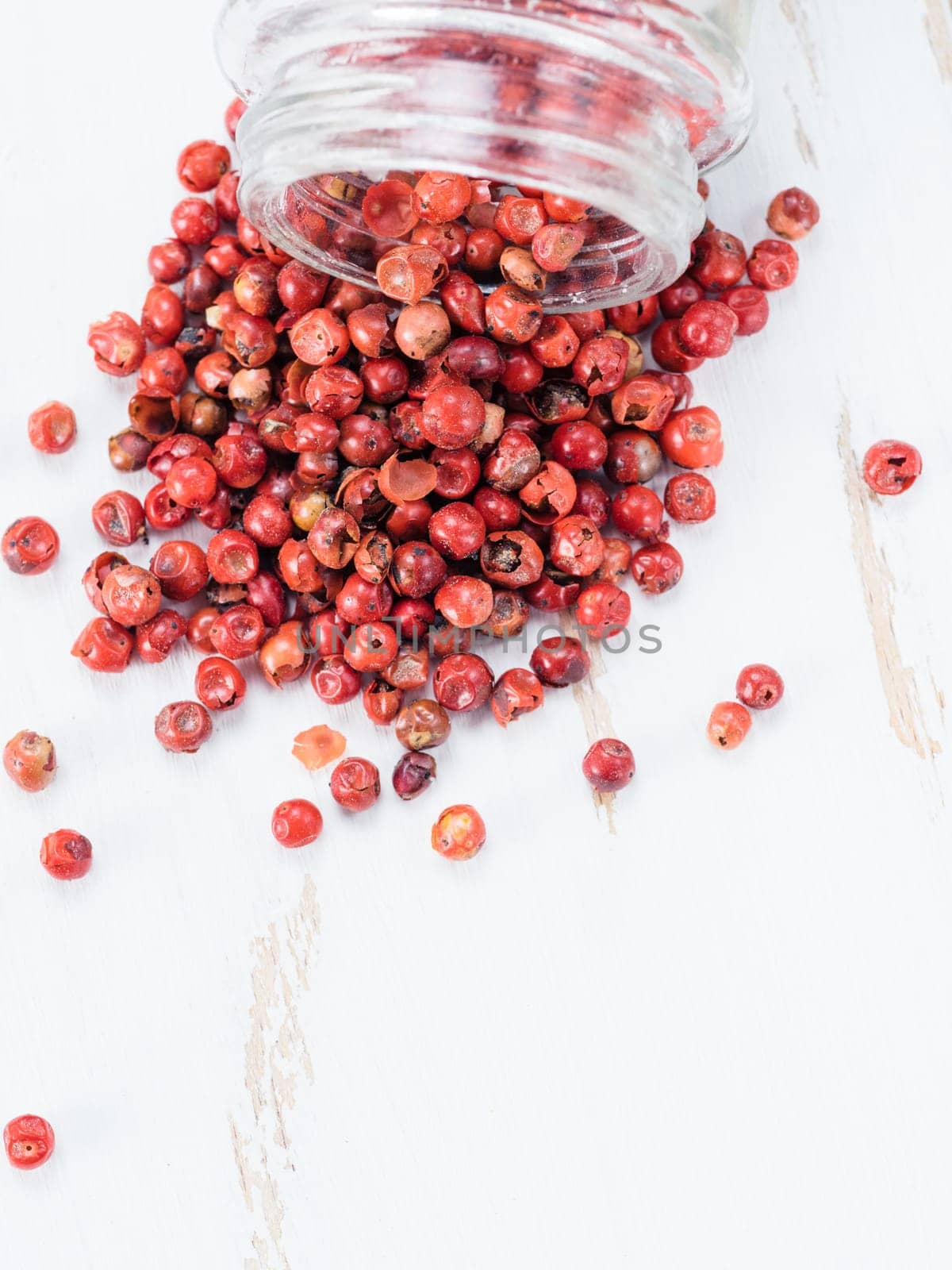 Dried pink pepper berries poured from glass jar on white wooden cutting board. Close up view of pink peppercorn poured on vintage wooden table. Copy space. Top view or flat-lay.