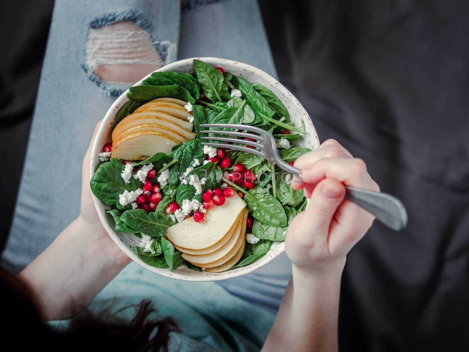 Woman in jeans holding vegan salad bowl with spinach, pear, pomegranate, cheese. Vegan breakfast, vegetarian food, diet concept. Girl in jeans holding fork with knees and hands visible