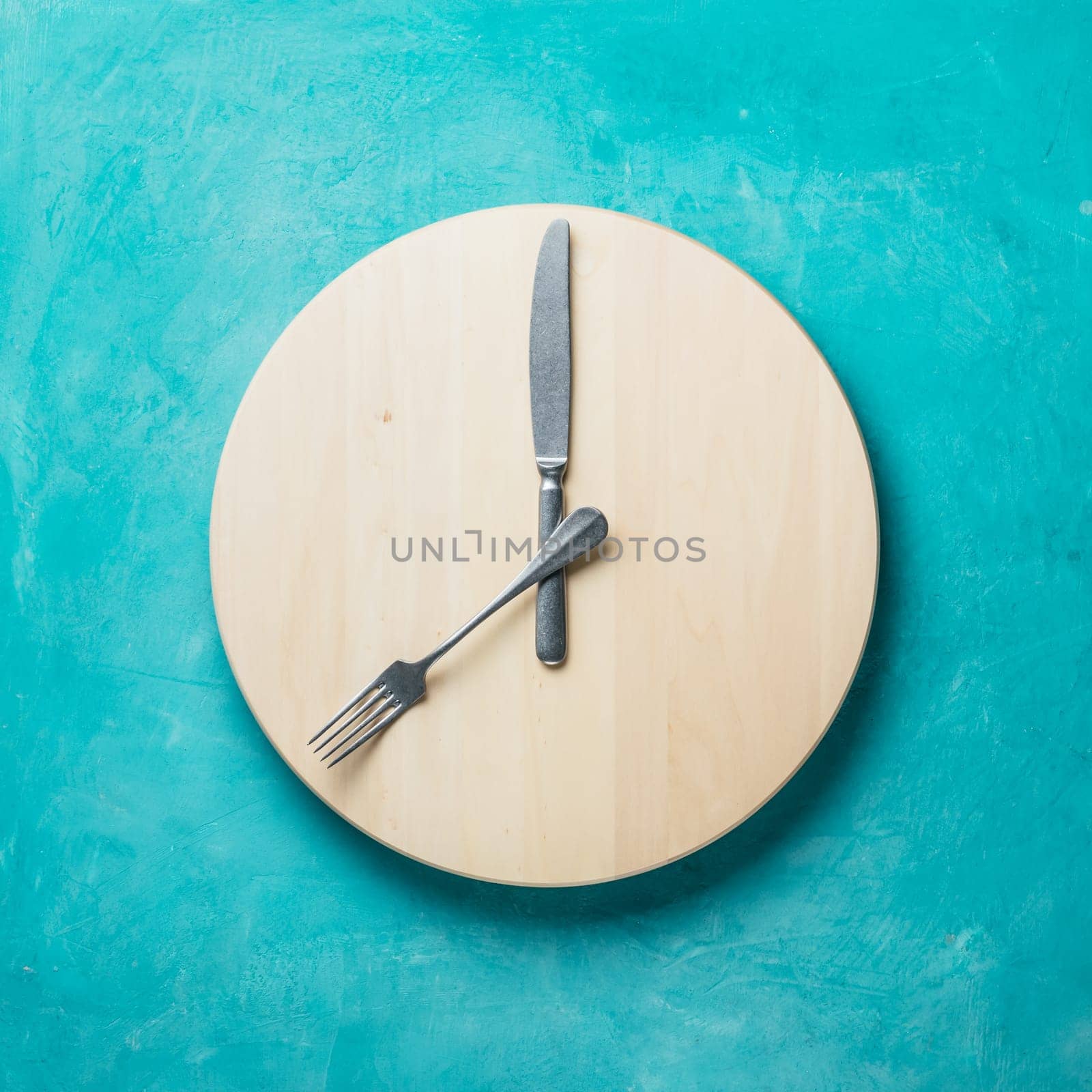 Intermittent fasting and skip breakfast concept - empty wooden round tray or trencher with cutlery as clock hands on blue background. Eight hour feeding window concept or breakfast time concept
