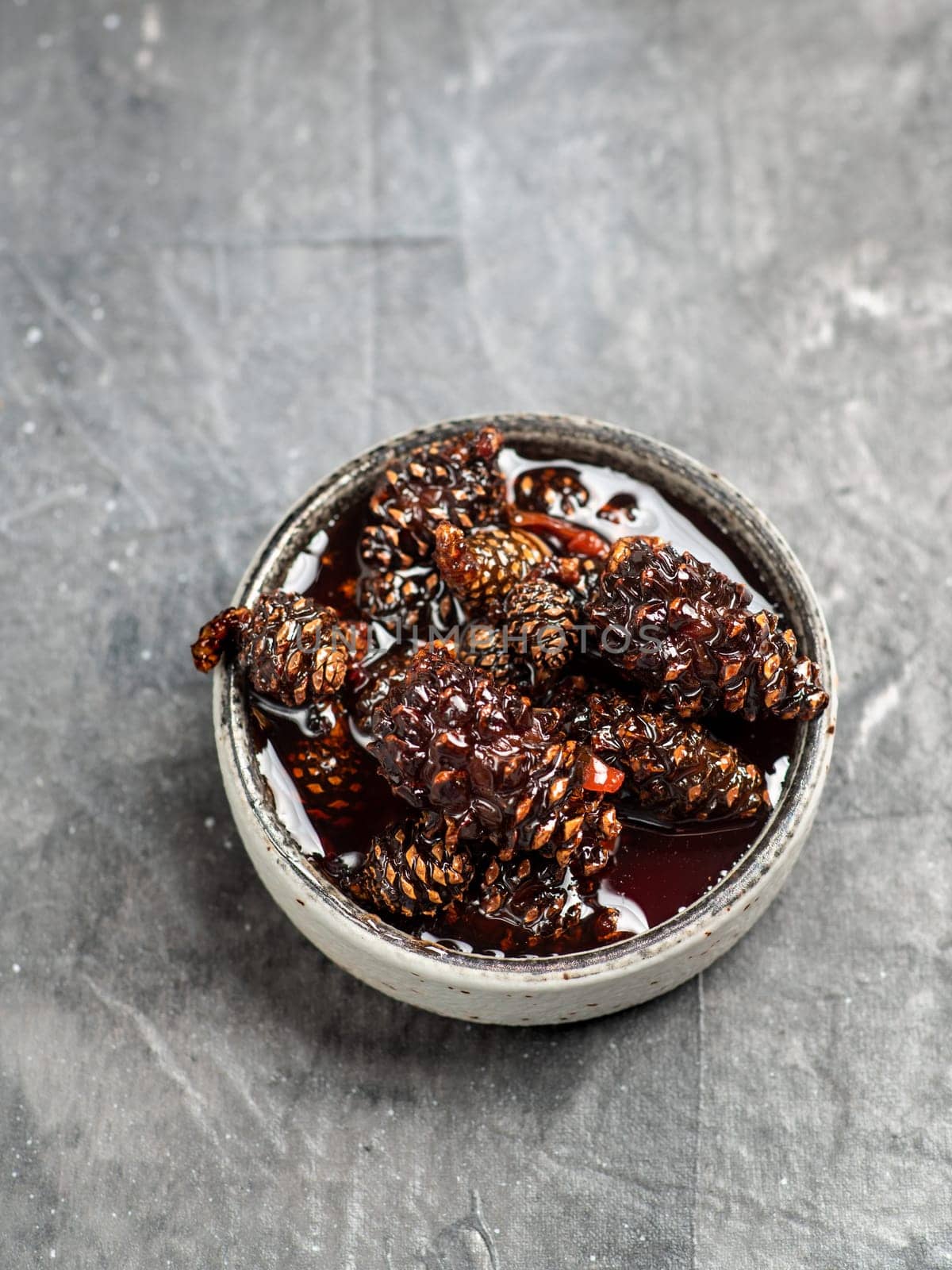 Delicious jam with baby pine cones in small bowl. Traditional Siberian dessert - young pine cones jam on gray textured background. Top view or flat lay.