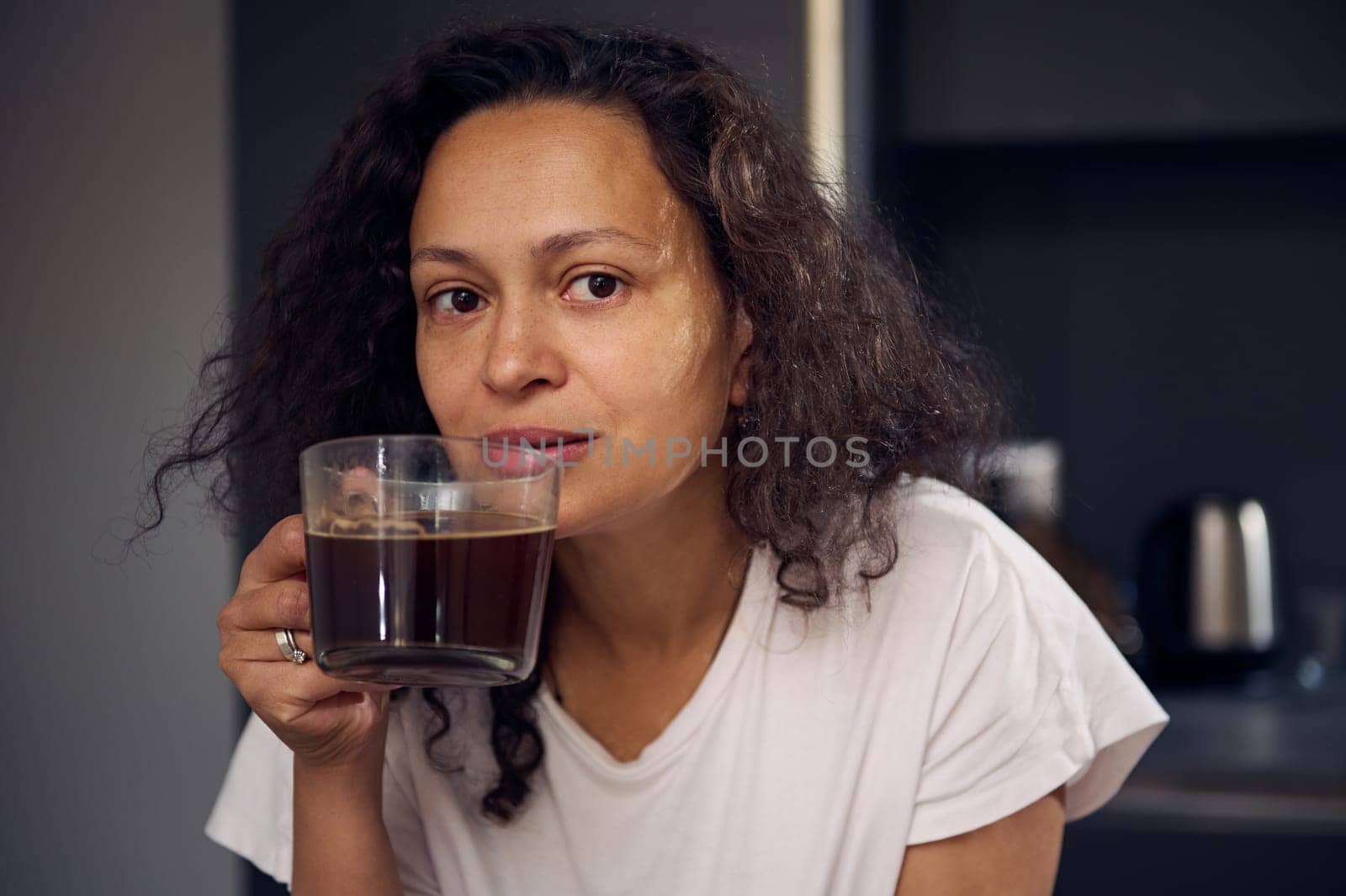 Beautiful woman holding glass cup of freshly brewed americano, smiling looking at camera. Curly haired woman in white pajamas, enjoying her morning espresso coffee. Morning routine. People. Lifestyle by artgf
