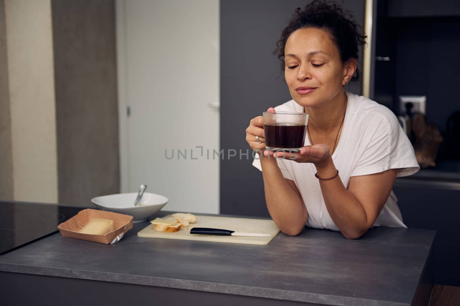 Confident multi ethnic curly haired brunette in white pajamas, holding a cup of espresso, smiling, standing with her eyes closed in the minimalist home kitchen interior, enjoying her morning coffee by artgf