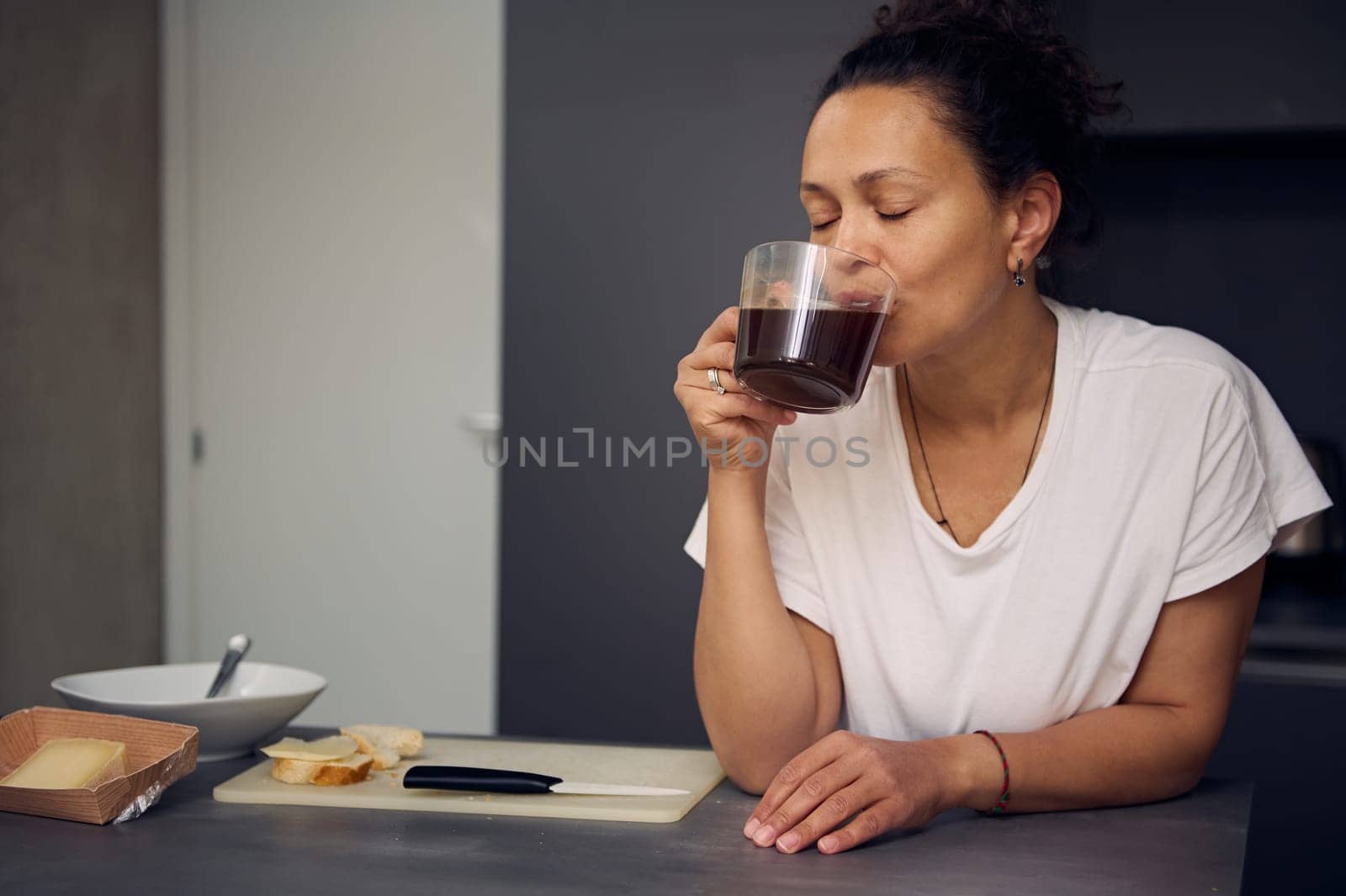 Portrait of relaxed happy woman in pajamas, taking a sip of freshly brewed espresso coffee during her breakfast in the morning, in minimalist home kitchen interior. People. Food and drink consumerism
