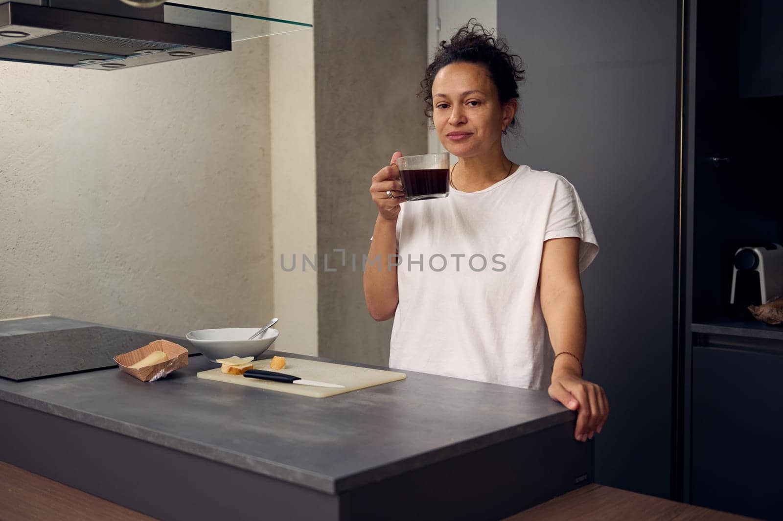 Beautiful multi ethnic positive woman enjoys her morning coffee, smiling looking at camera, standing at the kitchen table