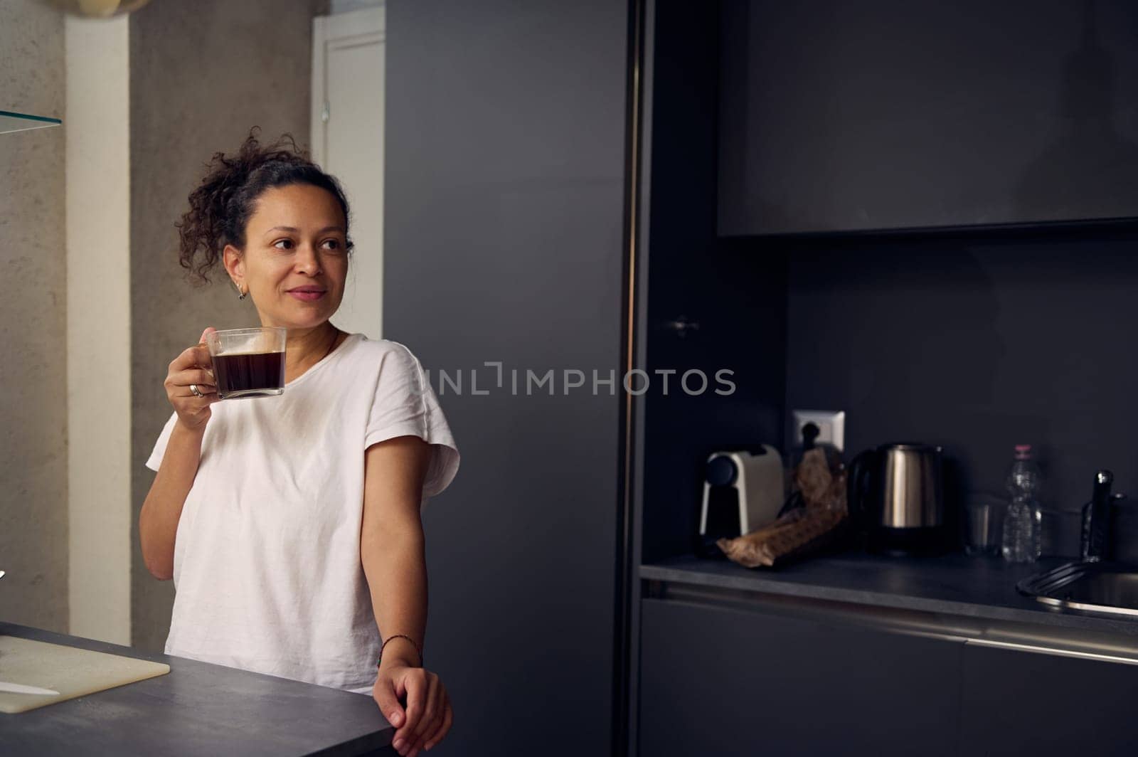 Multi ethnic pretty woman 40 years old, dressed in white pajamas, sipping a coffee for an energizing morning. Beautiful young woman drinking freshly brewed coffee at home, smiling looking away. by artgf