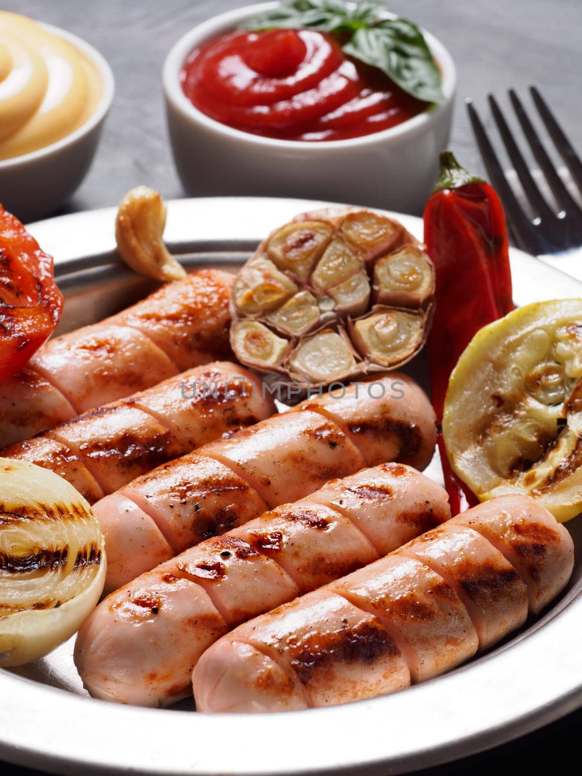 Chicken homemade sausages with sauces ketchup and mustard on black background. Grilled sausages and grilled vegetables in metal plate. Copy space. Vertical.