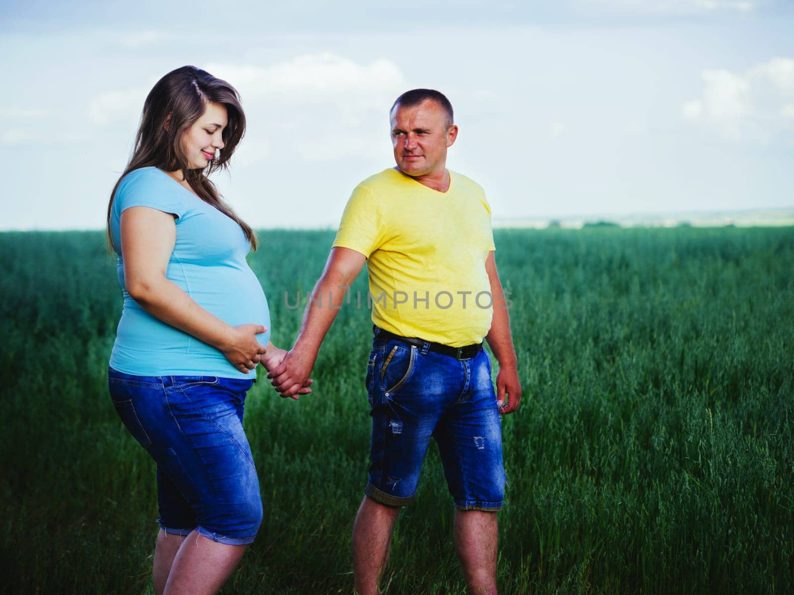 Smiling beautiful pregnant woman looking at her belly and holds hand of her husband outdoors. Casual couple holding hands and walking in a meadow.