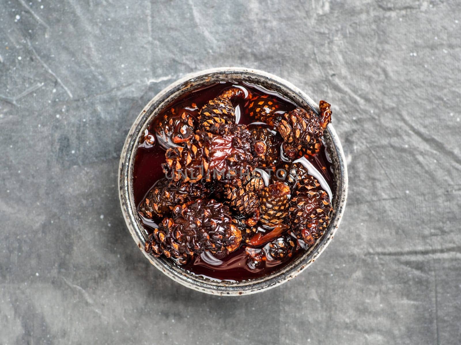 Delicious jam with baby pine cones in small bowl. Traditional Siberian dessert - young pine cones jam on gray textured background. Top view or flat lay.