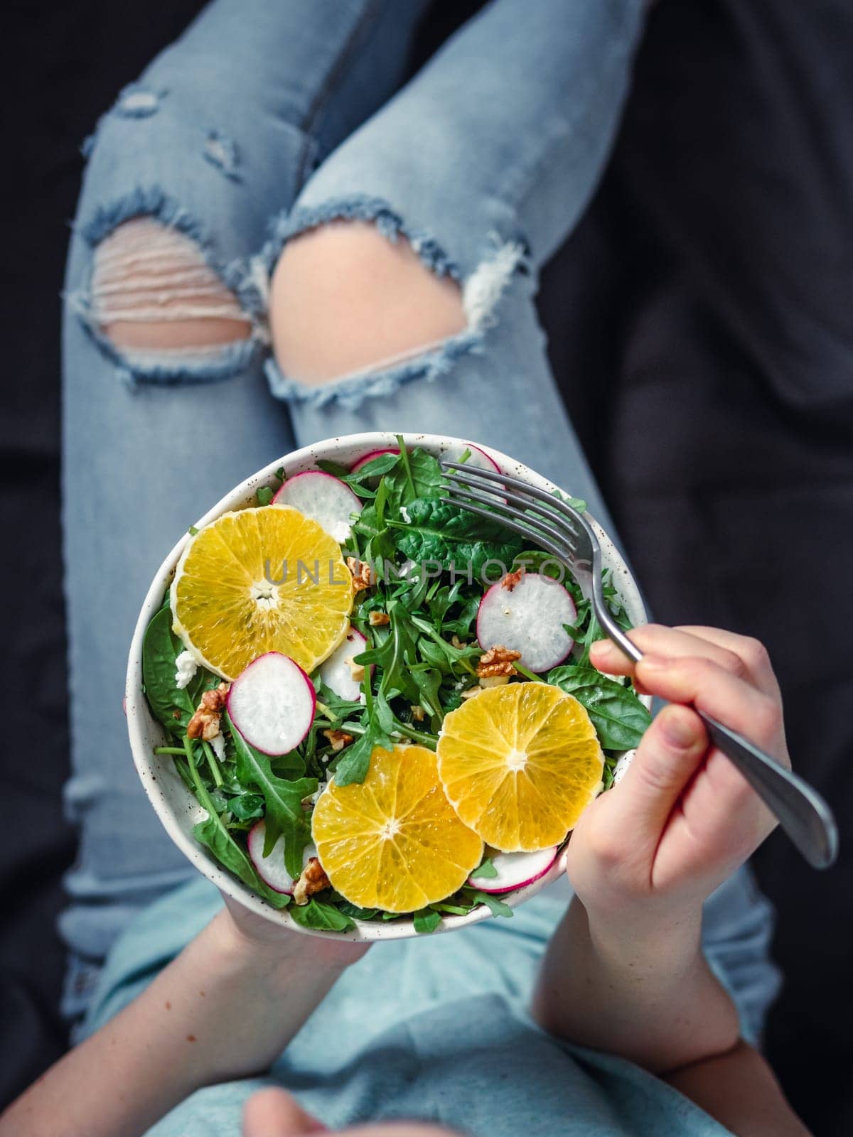 Woman in jeans at bed, holding vegan salad bowl with oranges, spinach,arugula,radish,nut.Top view.Vegan breakfast,vegetarian food,diet concept.Girl in jeans holding fork with knees and hands visible