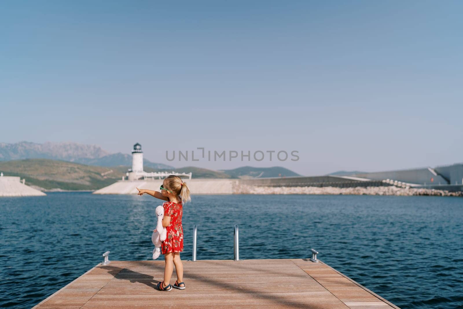 Little girl with a toy rabbit stands on the pier and points to the mountains. Back view. High quality photo