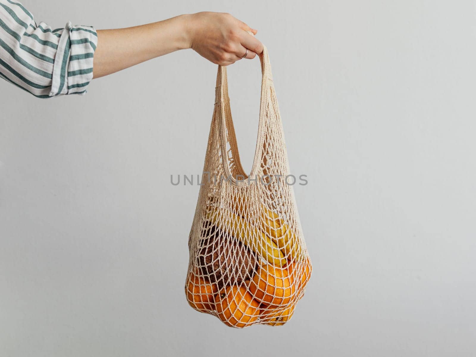 Mesh bag with fruits in female hand. Stylish young woman hand hold mesh shopping bag on light gray wall. Modern reusable shopping concept.