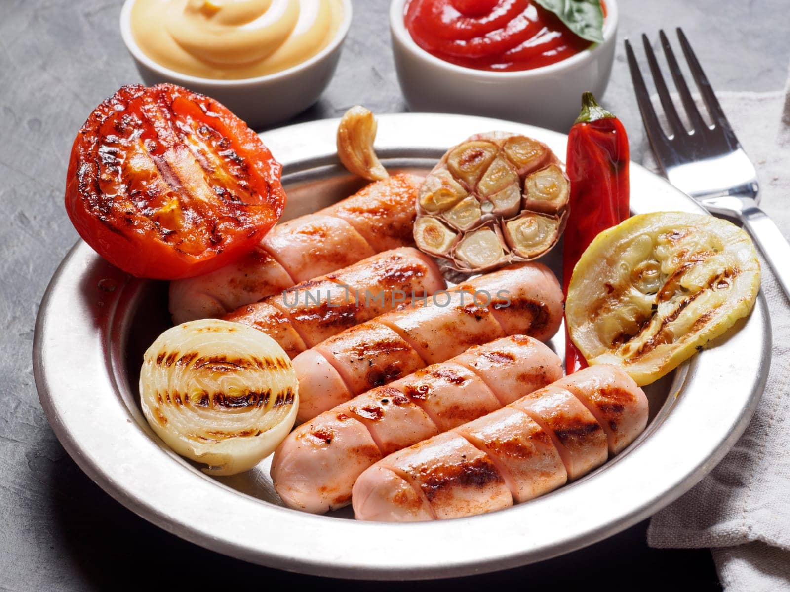 Chicken homemade sausages with sauces ketchup and mustard on black background. Grilled sausages and grilled vegetables in metal plate. Copy space.