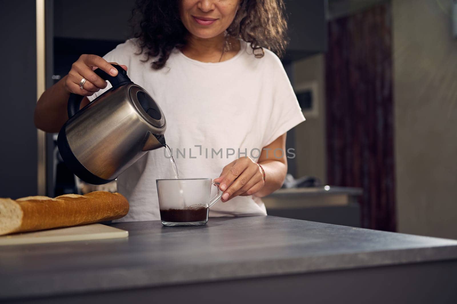 Close-up of a smiling young woman pouring some boiled water from an electric tea pot into a cup of soluble coffee, preparing her coffee drink in the morning by artgf