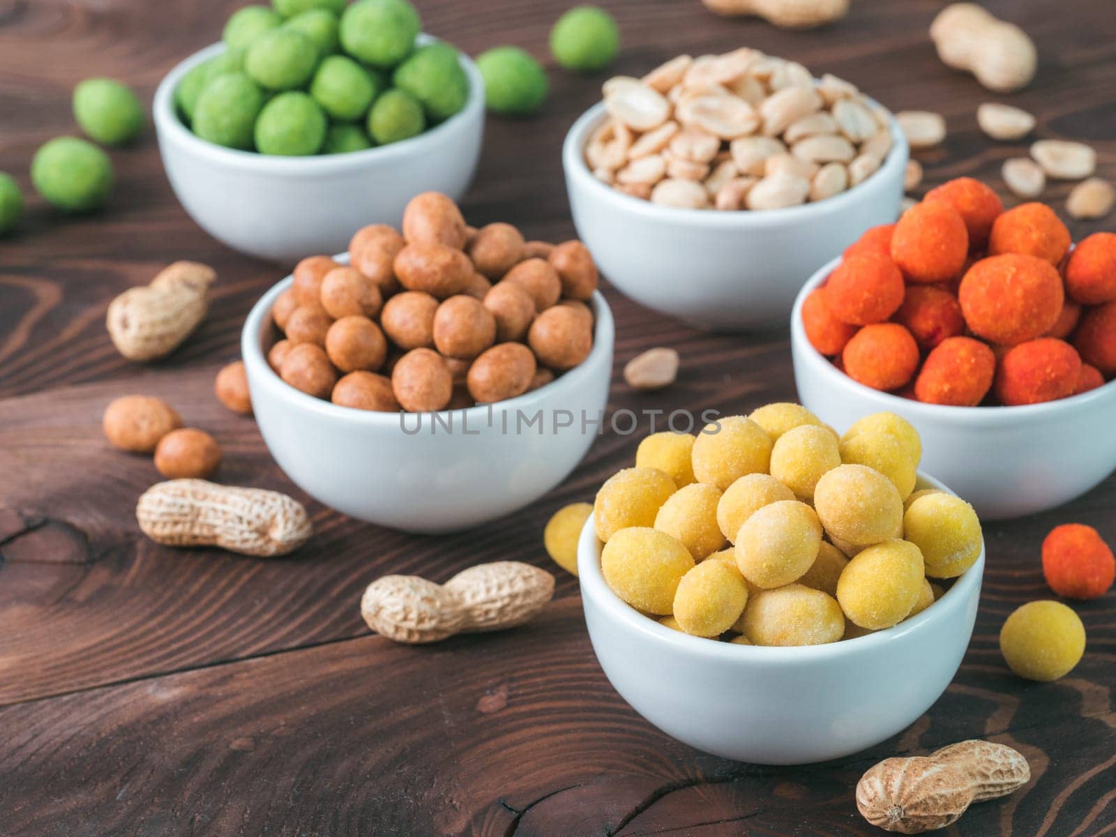 Assortment of different peanuts snack for beer, wine, party bar. Salted peanuts, peanuts in coconut glaze, green vasabi, red spicy chilli, yellow cheese glaze on brown wooden table. Copy space.