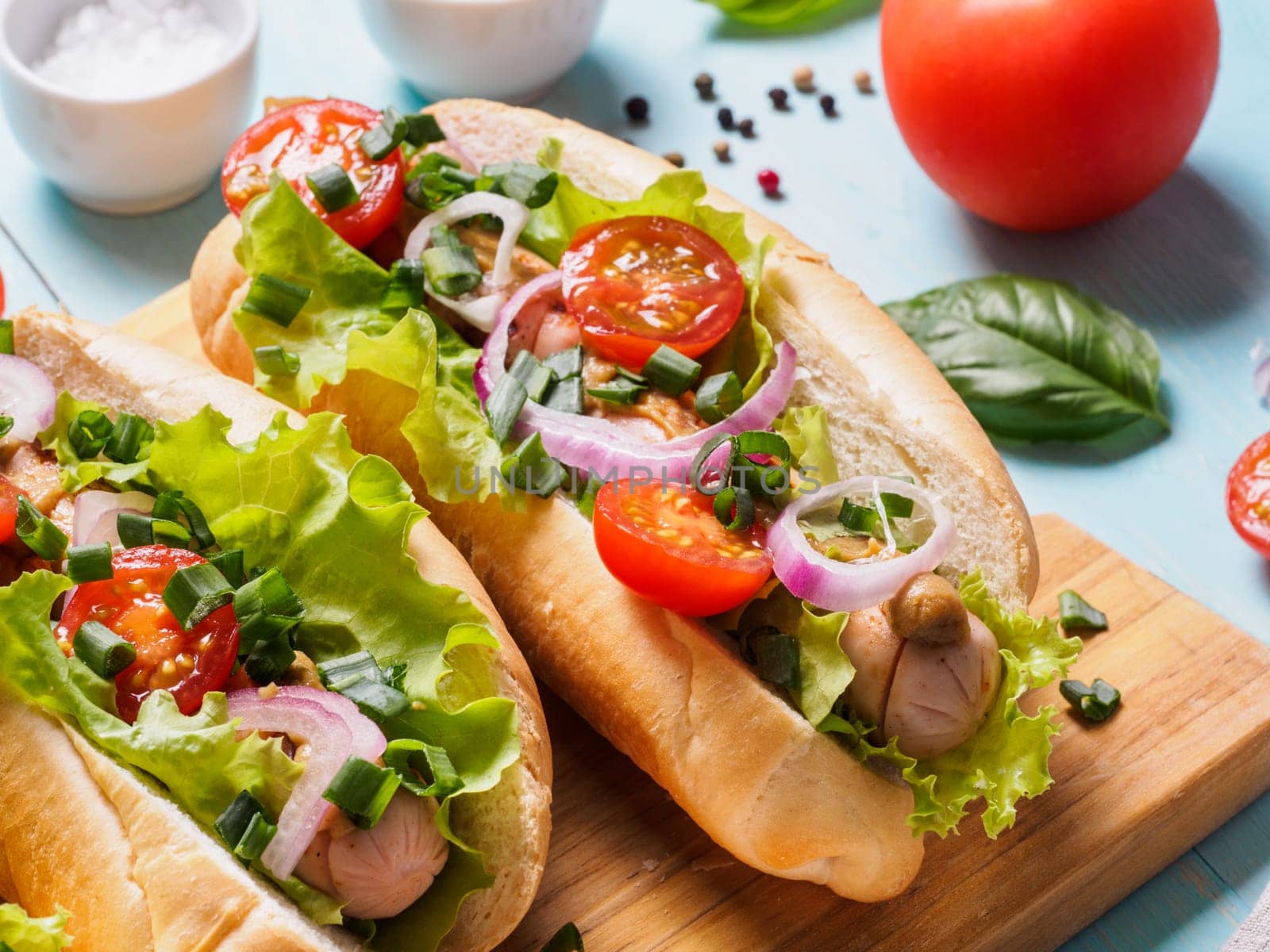 Close up view of homemade hotdogs with chicken sausages, fresh vegetables on blue wooden background. Hot-dogs with tomatoes, onion, basil, letucce and spices.