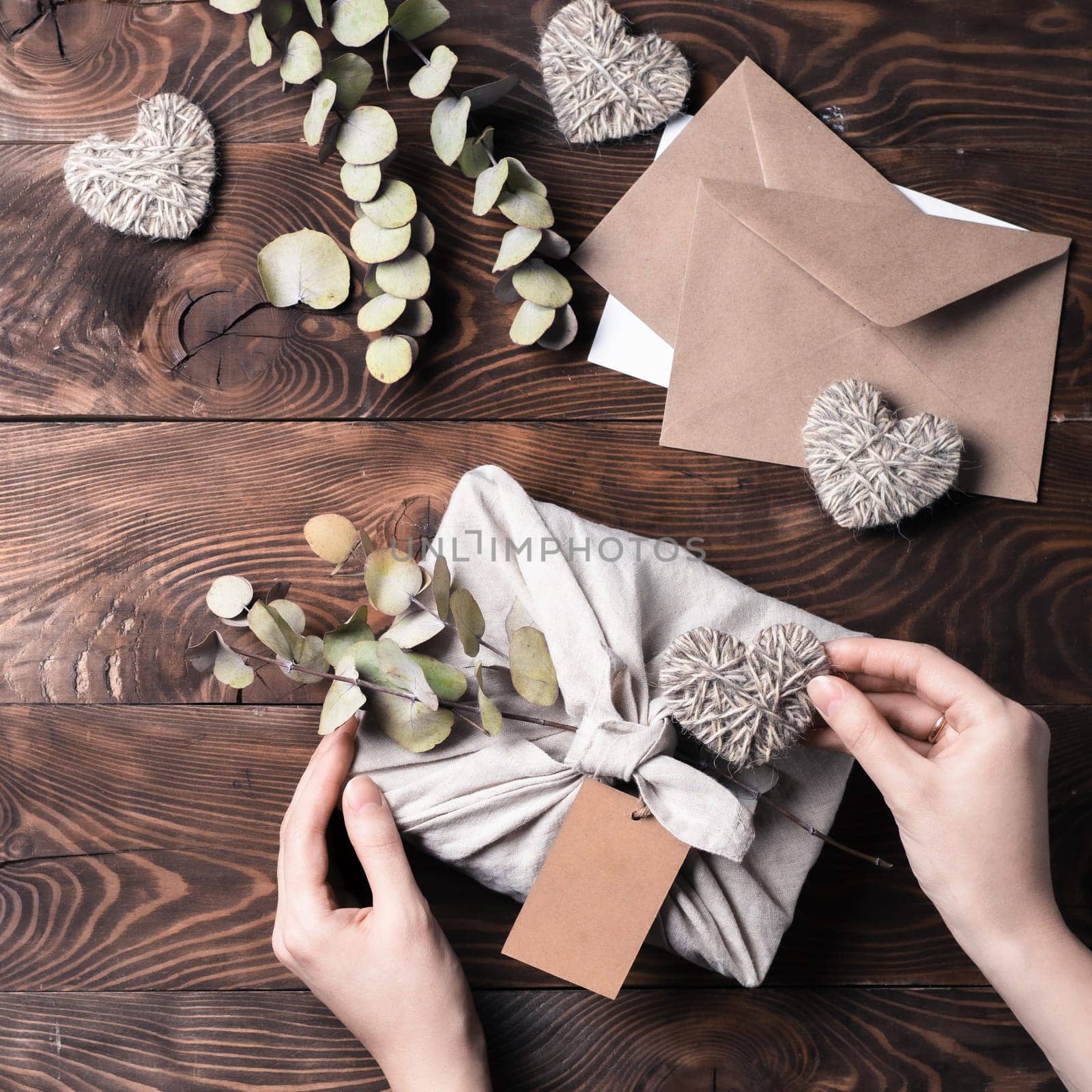 Zero waste, Valentine's Day eco-friendly gift wrapping in Furoshiki style with dry eucalypt. Hands with gift box in cloth wrapping with empty craft label on brown wooden background. Top view, flat lay