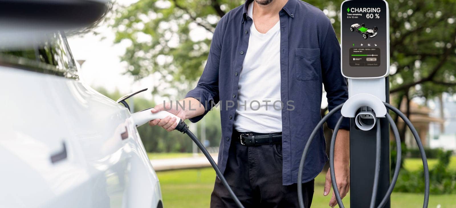 Man recharge EV electric vehicle's battery from EV charging station in outdoor green city park scenic. Eco friendly urban transport and commute with eco friendly EV car travel. Exalt