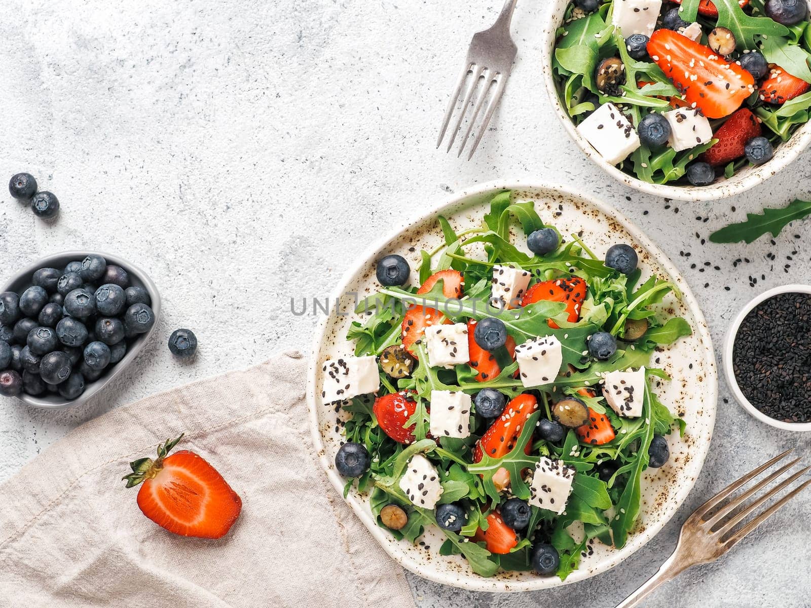 Salad with arugula, feta cheese and berries - strawberry, blueberry, in craft plate on gray cement bacground. Summer salad idea and recipe for healthy vegetarian lunch, dinner. Top view. Copy space.