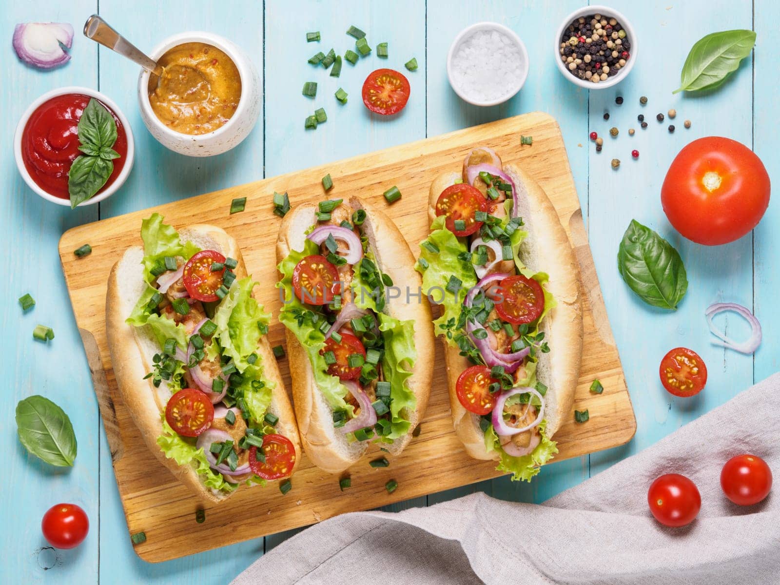 Top view of homemade hotdogs with chicken sausages, fresh vegetables, ketchup and mustard sauce on blue wooden background. Hot-dogs with tomatoes, onion, basil, letucce and spices.